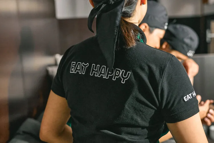 EatHappy Mitarbeiterin in schwarzer Arbeitskleidung mit Rückenaufdruck Eat Happy im Gespräch mit Kolleginnen und Kollegen