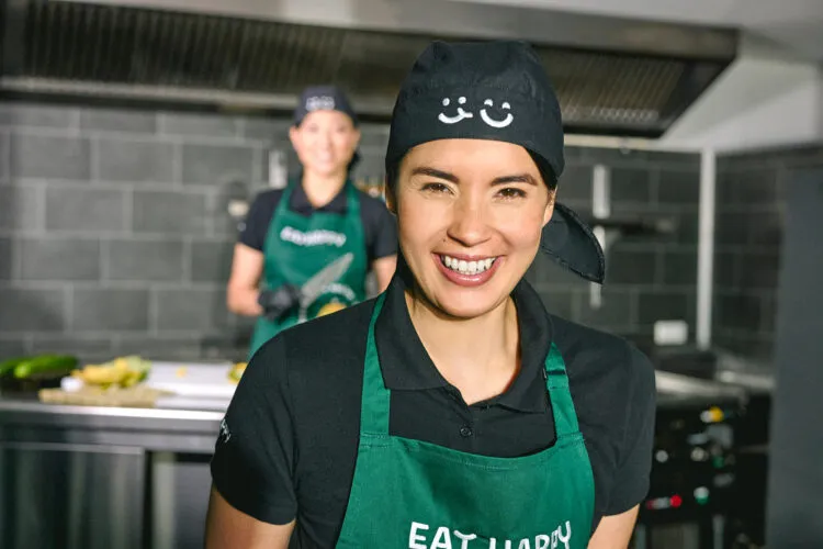 EatHappy Mitarbeiterin mit grünem Schürze und schwarzer Kochmütze lächelt in einer Küche