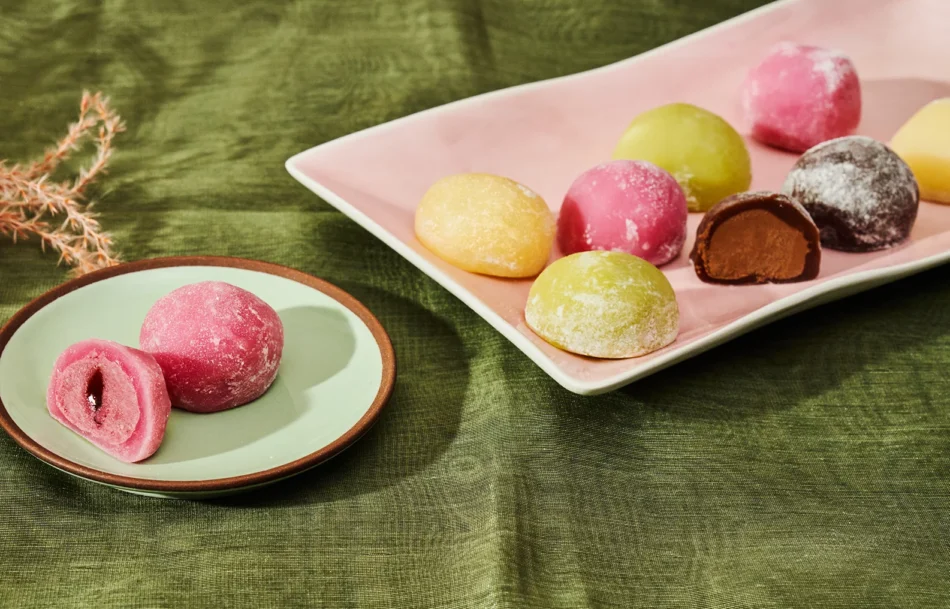 Bunte Mochi in Rosa, Gelb, Grün und Braun auf Tellern vor grünem Stoffhintergrund